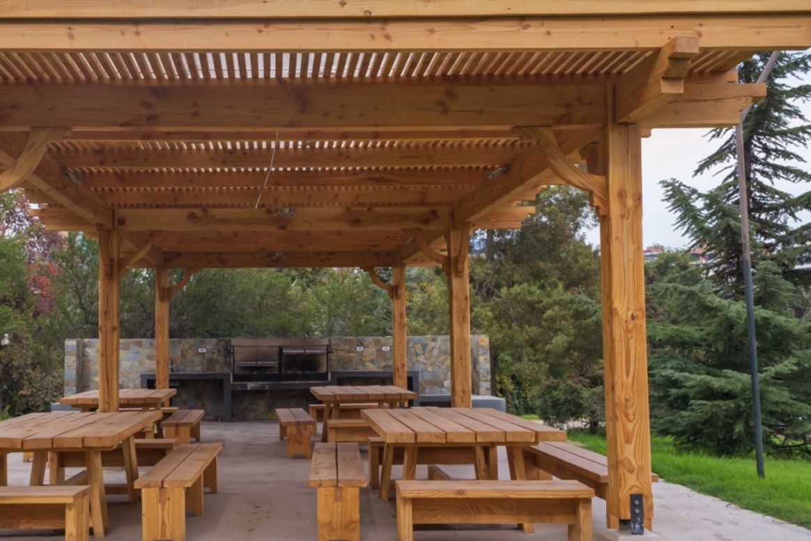 Quincho institucional con estructura de madera laminada, mesas y bancas de madera y parrilla de piedra al fondo