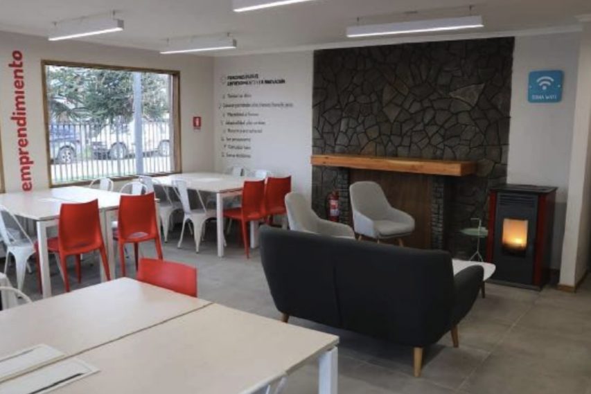 Espacio de cotrabajo con mesas blancas, sillas rojas, sofá gris y chimenea con muro de piedra